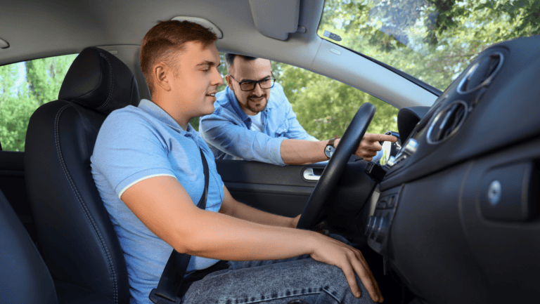 A young man sits in the driver’s seat as an instructor leans in from outside, guiding him through the dashboard controls during driver onboarding.