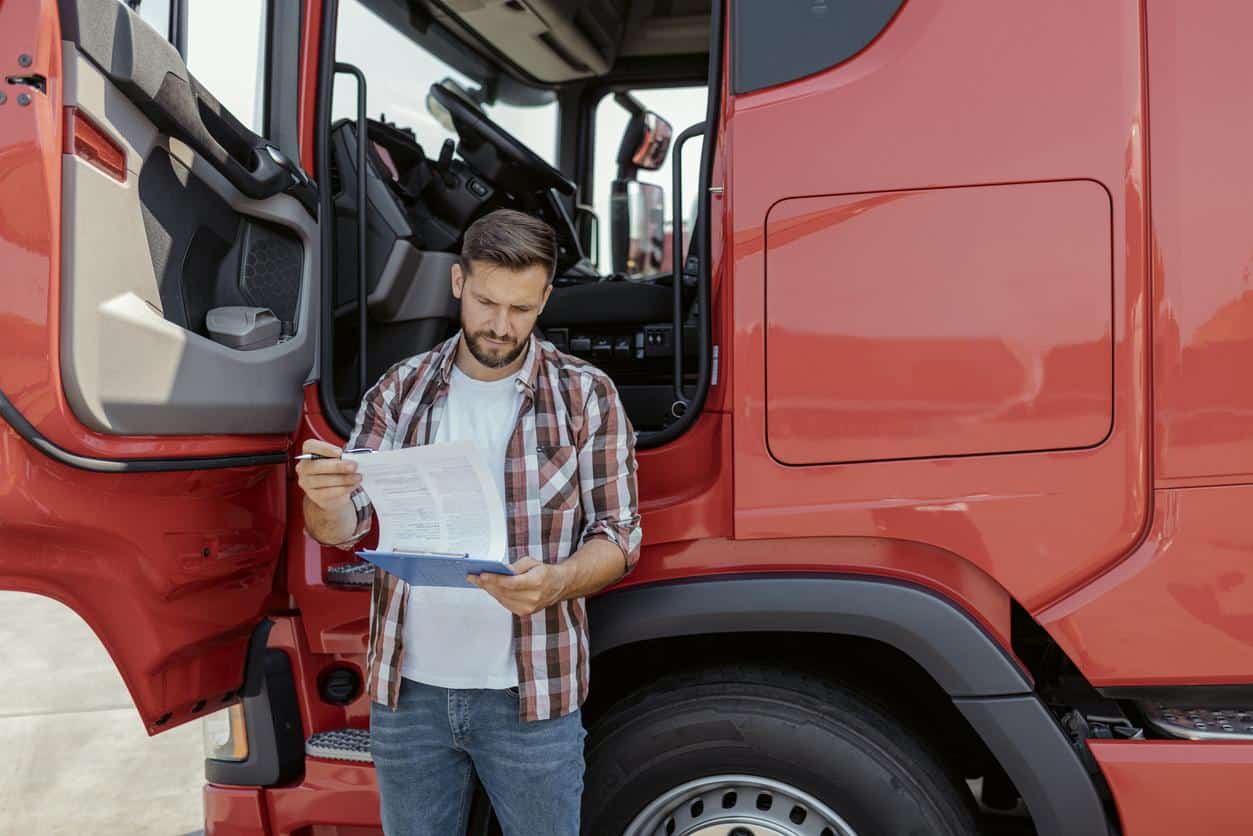 Man standing beside a red truck, holding a clipboard and pen, reviewing paperwork near the open drivers side door.